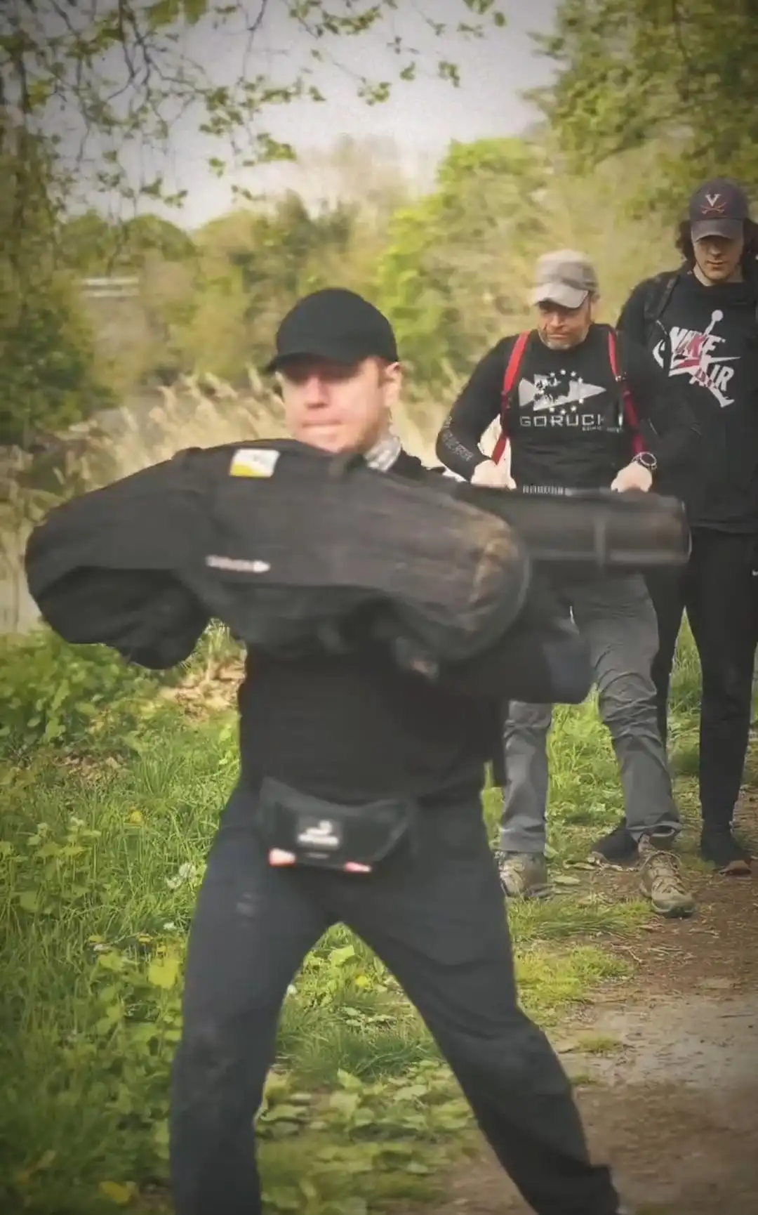 Image: Image view is front-to-back during a group exercise, centered on a participant's effort to throw a sandbag forward, captured mid-movement, sandbag in top position