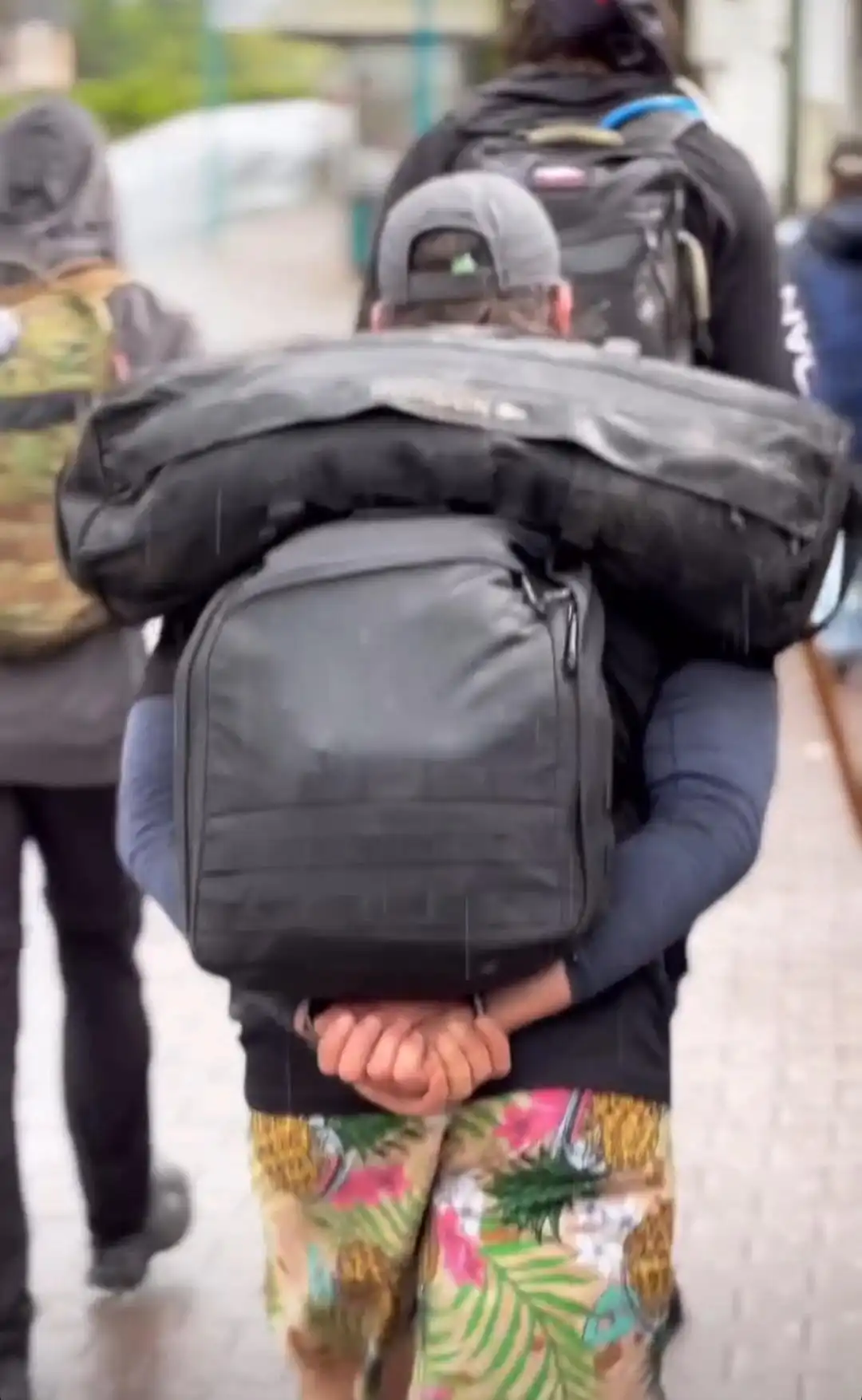Image: View from the back of a group movement through the city. Close-up on a participant with a sandbag on top of his ruck. Below the ruck, we can see his hands are folded for extra support