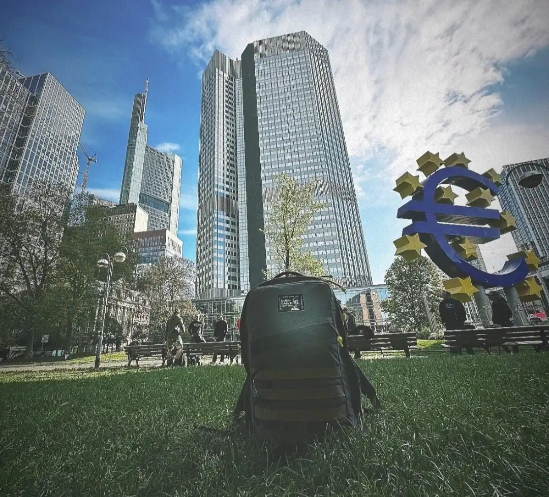 Image: Image taken from a frog perspective of a ruck sitting on the grass in front of the Euro Sculpture. The ruck displays a commemorative patch for Milton Davison. Benches, buildings and people can be seen in the background. 